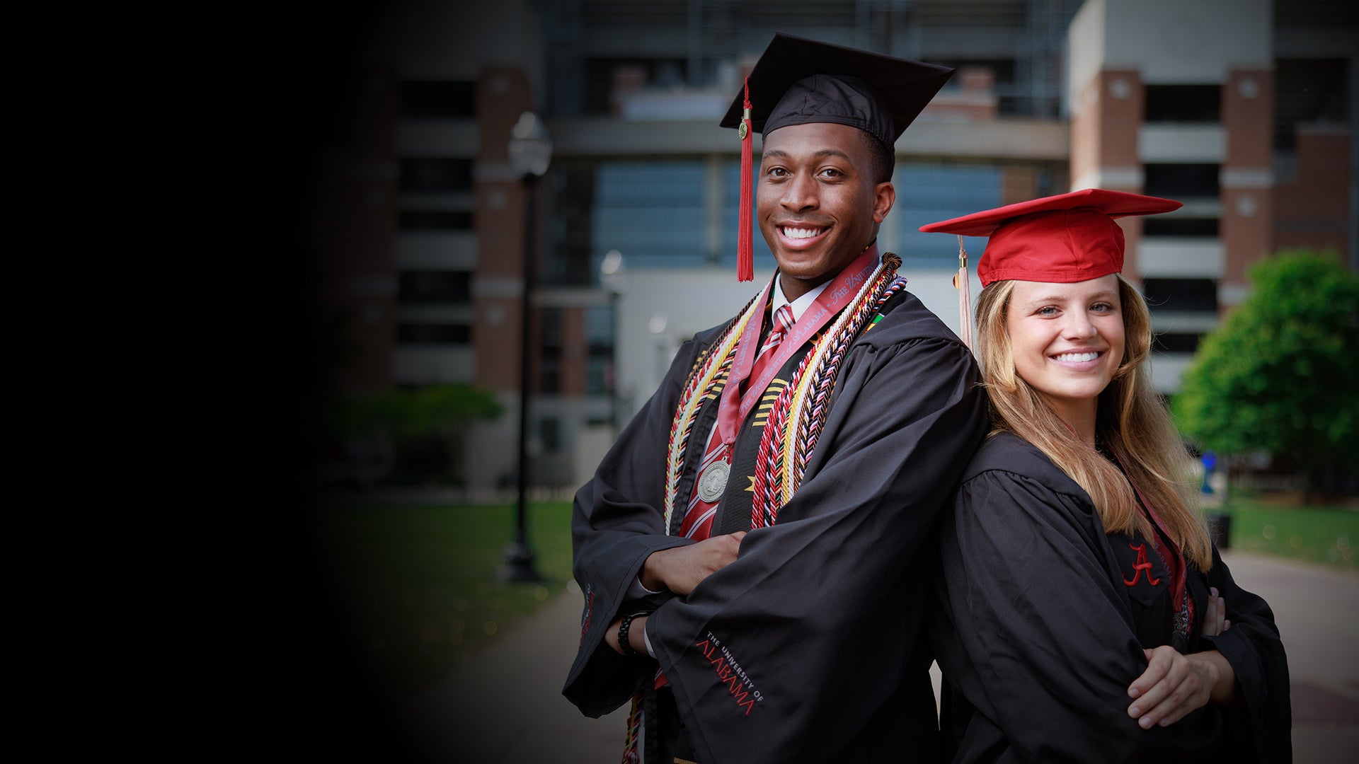Two students posing in graduation regalia in front of Bryant-Denny Stadium